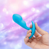 A hand holds the B-Vibe Vibrating P-Spot Snug Plug RC Small by B-Vibe, a blue prostate stimulator, against a blurred pastel purple and pink background with bokeh lights, emphasizing its design for targeted stimulation.